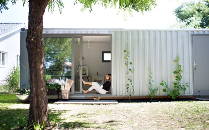 Femme mature adossé à la porte coulissante d'une maison container posée sur un terrain et aménagée.